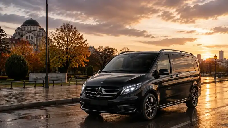 Crni Mercedes kombi za prevoz putnika na mokrom platou u zalasku sunca, sa kupolom pravoslavnog hrama i jesenjim drvoredom u pozadini